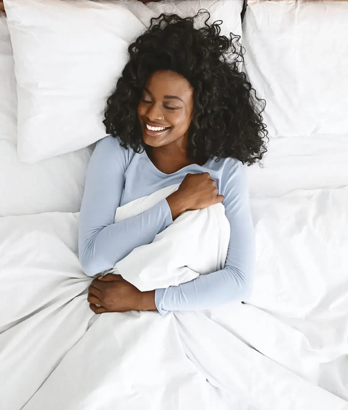 Lachende vrouw ligt in bed onder een wit dekbed.
