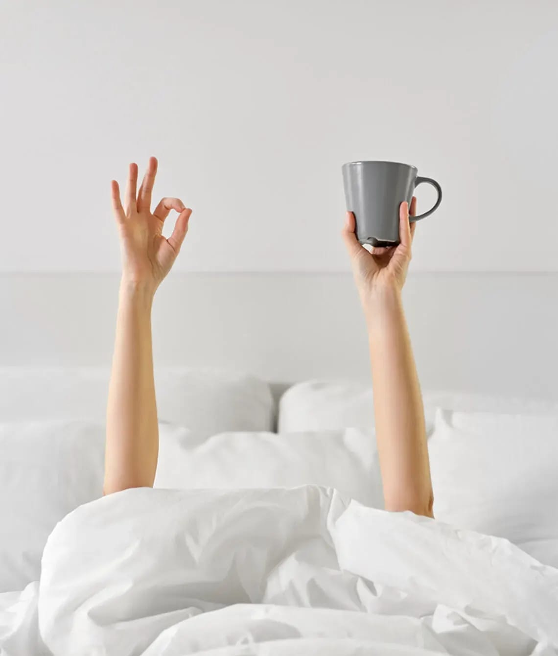 Armen met een kop koffie steken boven een wit dekbed uit.