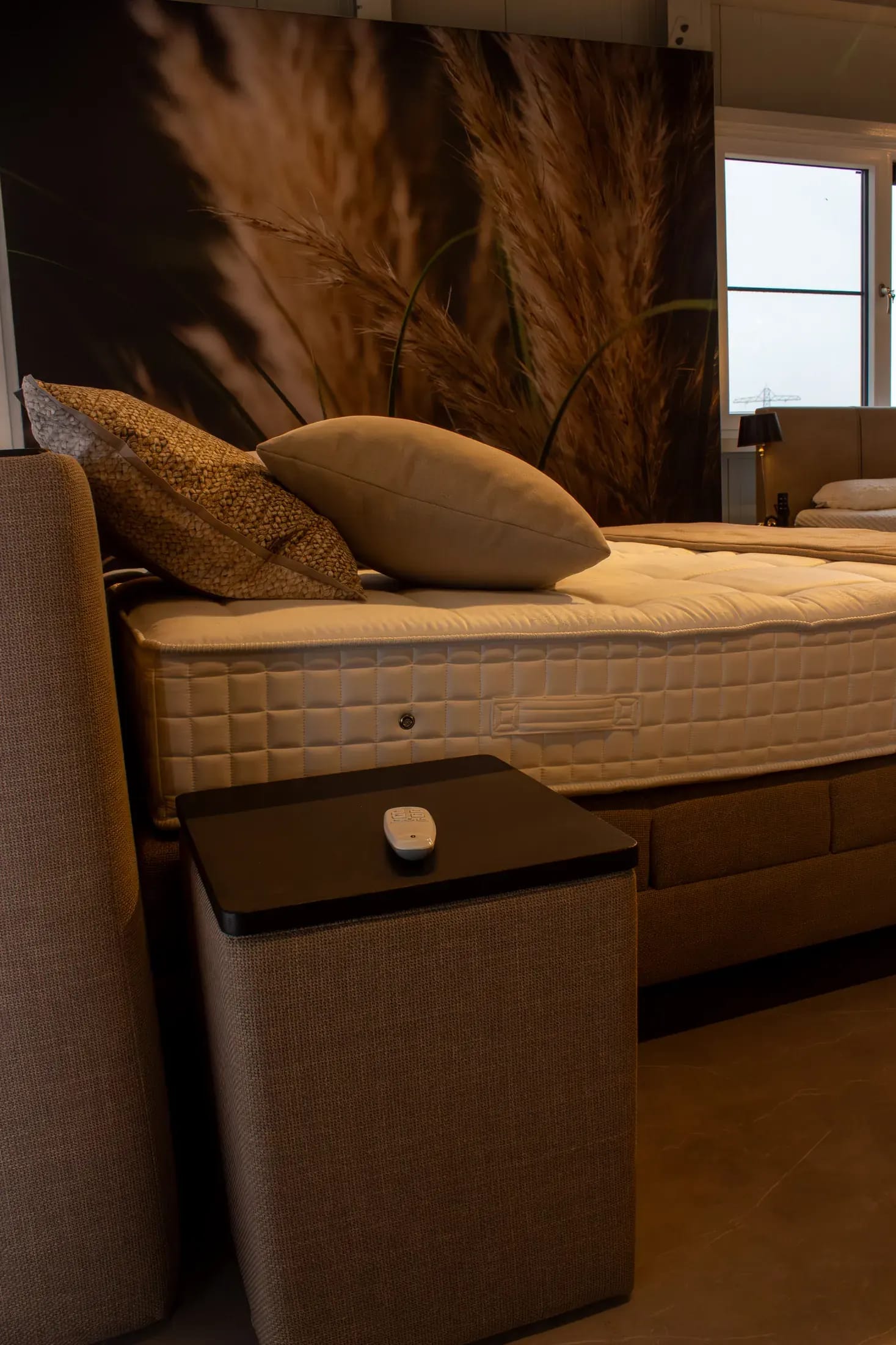 Close-up van een boxspring met gestoffeerd hoofdbord, sierkussens en een nachtkastje met afstandsbediening.