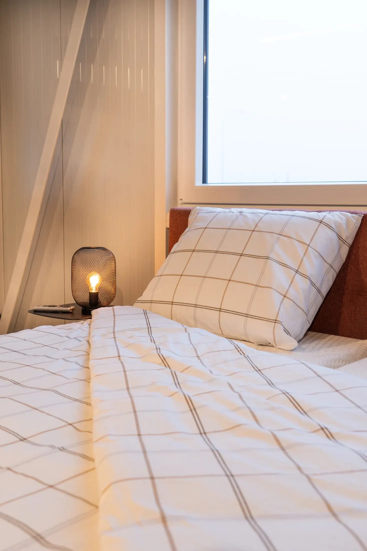 Boxspring met geruit beddengoed en een sfeerlamp bij het raam.