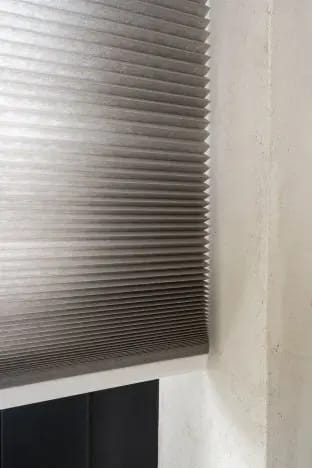 Taupe honingraatgordijn voor een raam naast een betonnen wand.