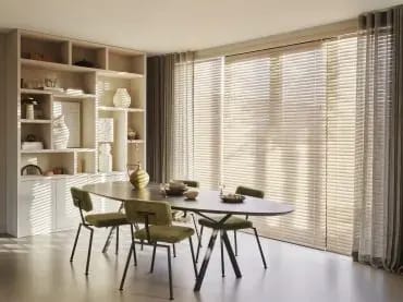 Houten jaloezieën voor de ramen van een eetkamer met ovale tafel.