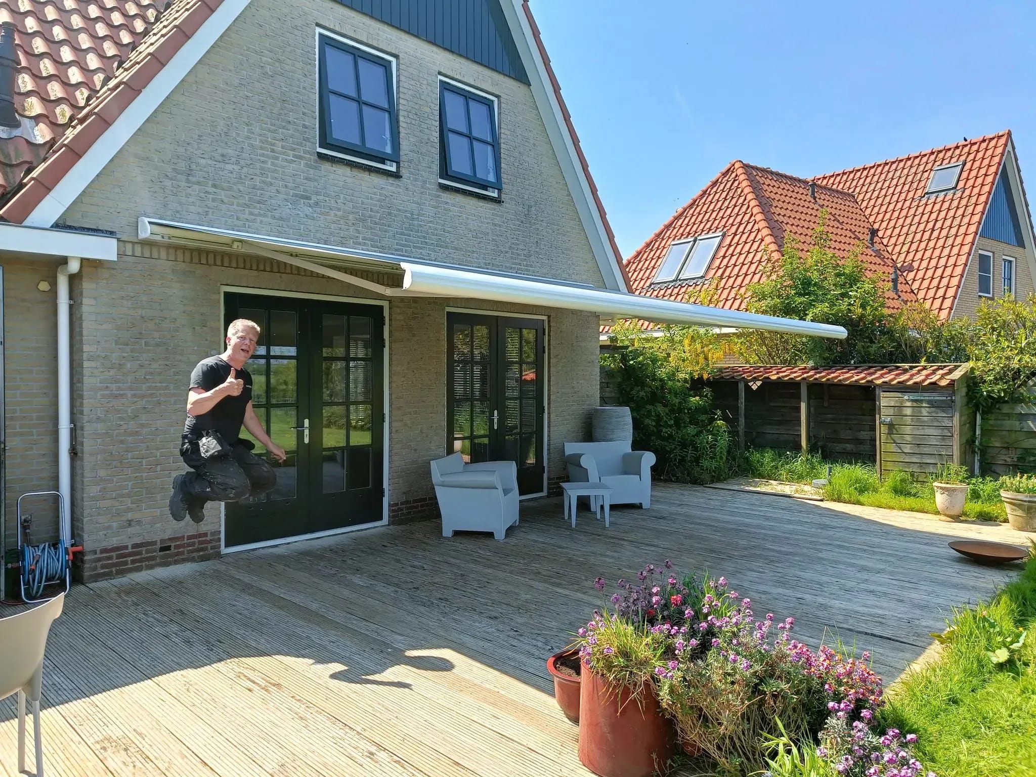 Uitgeklapt knikarmscherm boven een terras; de vakman springt blij de lucht in.