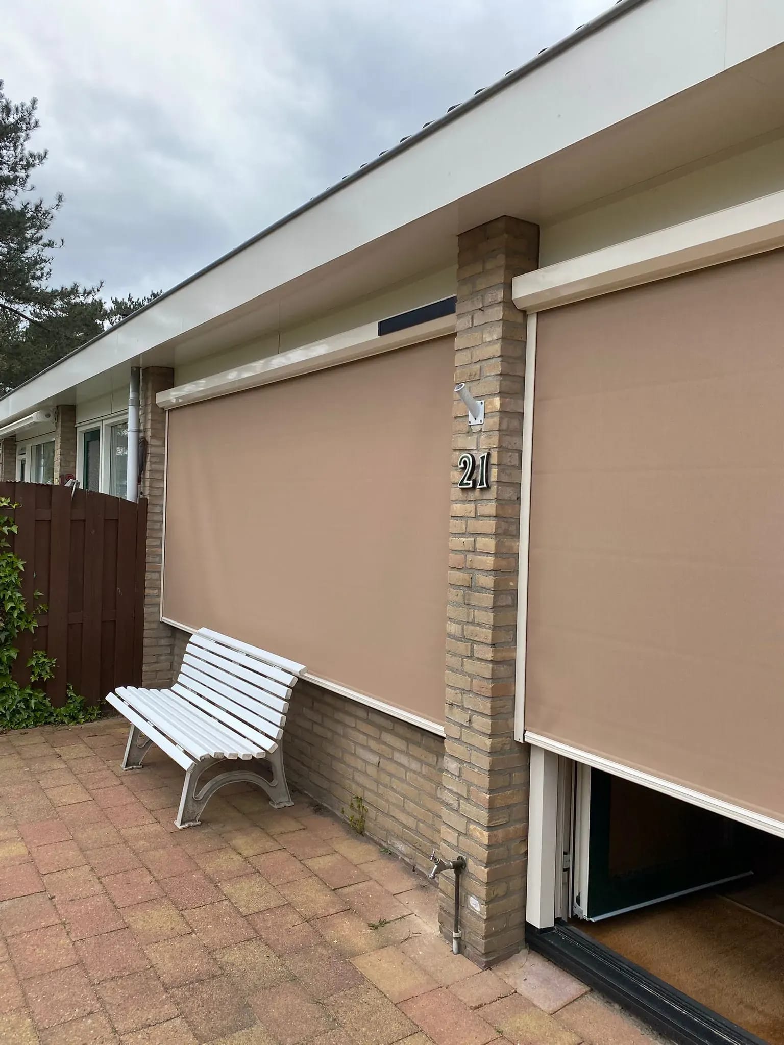 Uitgerolde zandkleurige screens voor de tuindeuren, met een wit bankje ervoor.