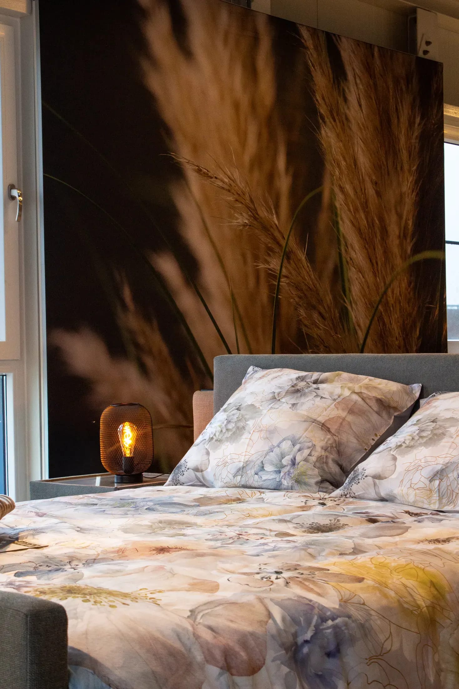 Bed met een dekbedovertrek met bloemmotief en een sfeerlamp, voor een wand met pampasgras.
