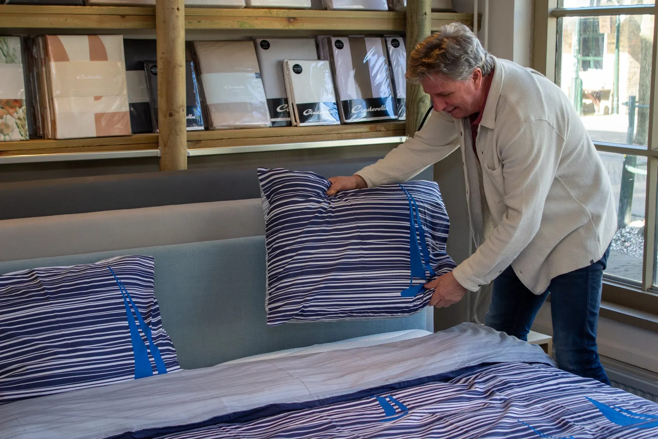 Medewerker schudt een blauw gestreept kussen op, met pakken beddengoed op de plank erachter.