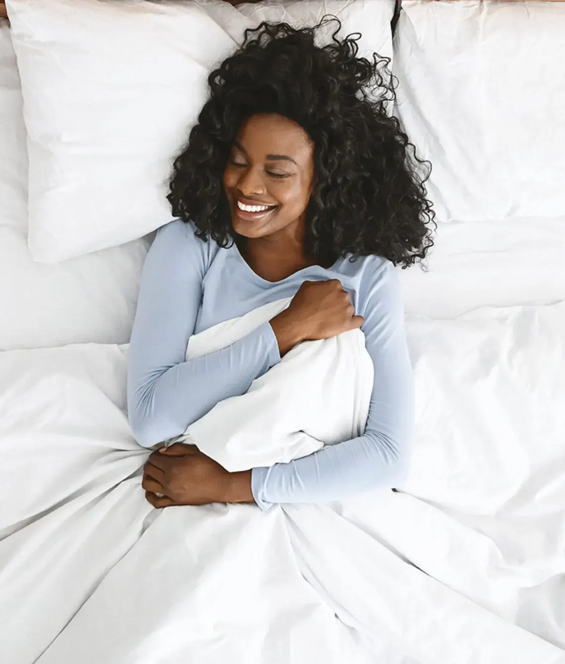 Lachende vrouw ligt in bed onder een wit dekbed.