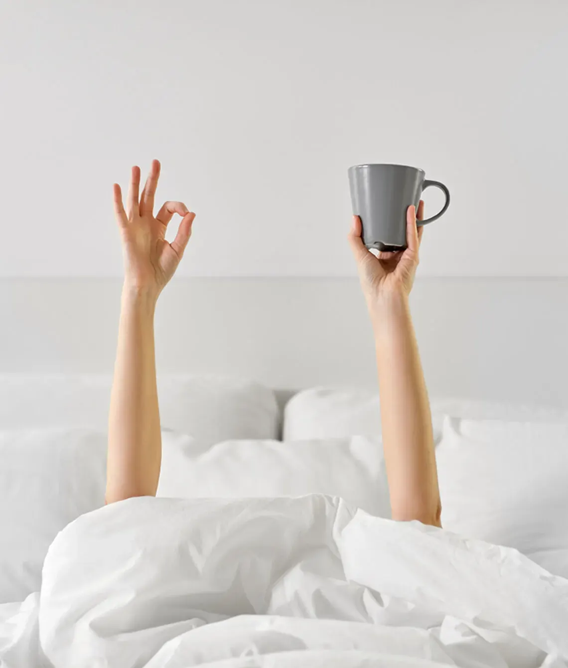 Armen met een kop koffie steken boven een wit dekbed uit.