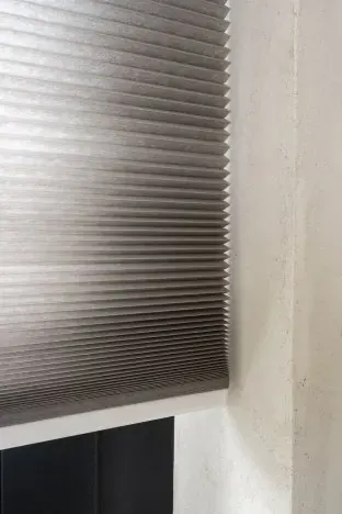 Taupe honingraatgordijn voor een raam naast een betonnen wand.