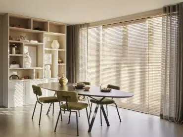 Houten jaloezieën voor de ramen van een eetkamer met ovale tafel.