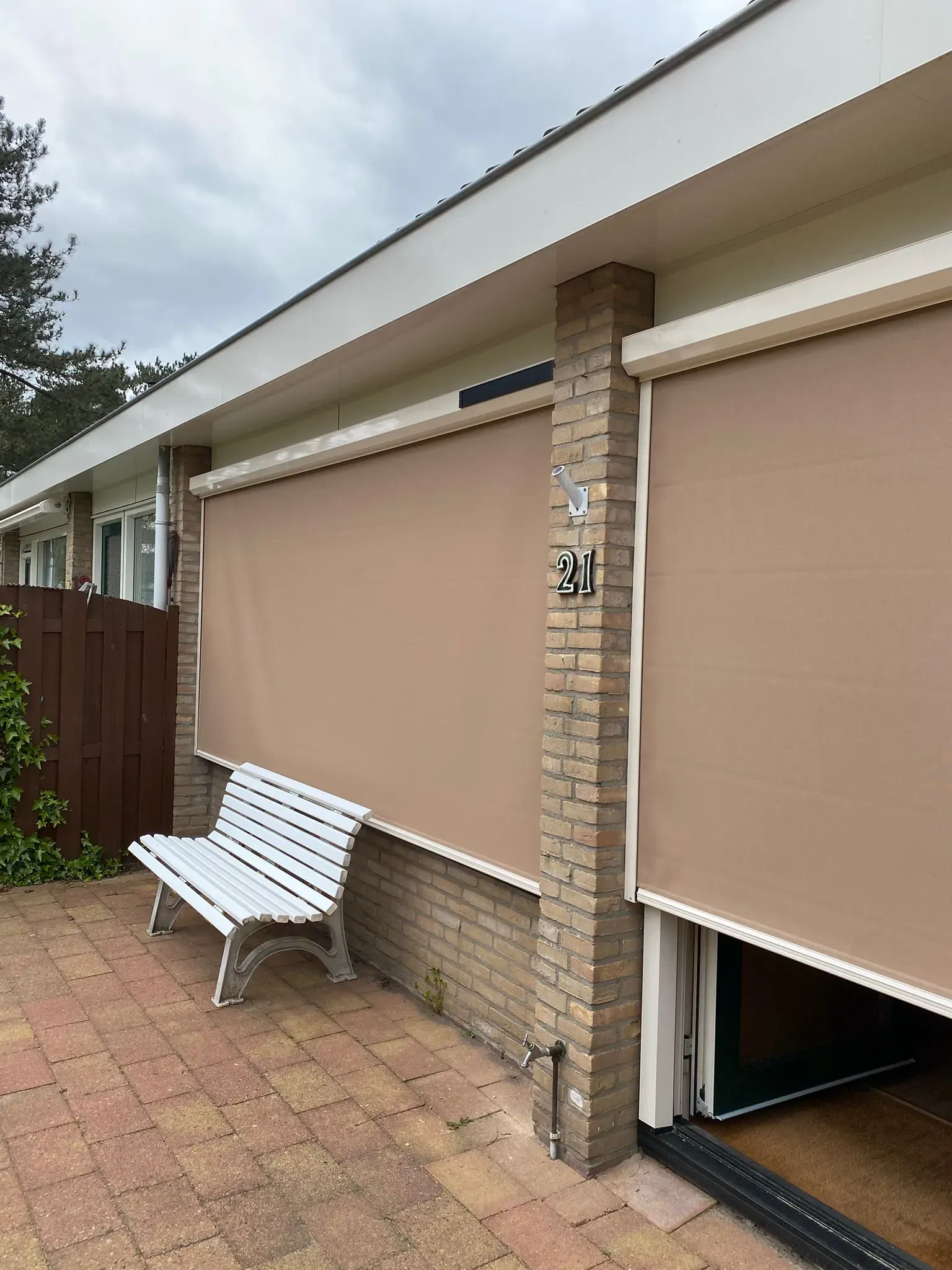 Uitgerolde zandkleurige screens voor de tuindeuren, met een wit bankje ervoor.