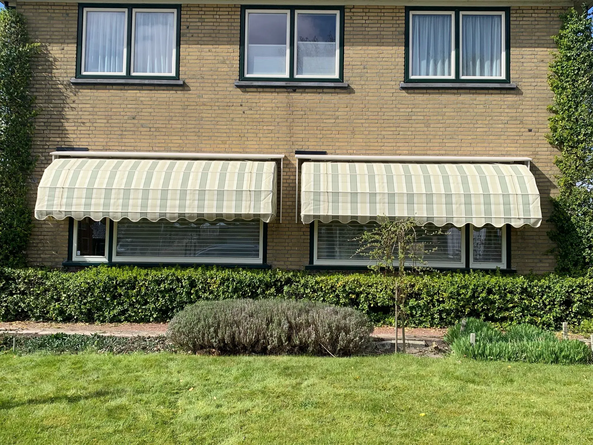 Twee geel-wit gestreepte markiezen boven de benedenramen van een woning.